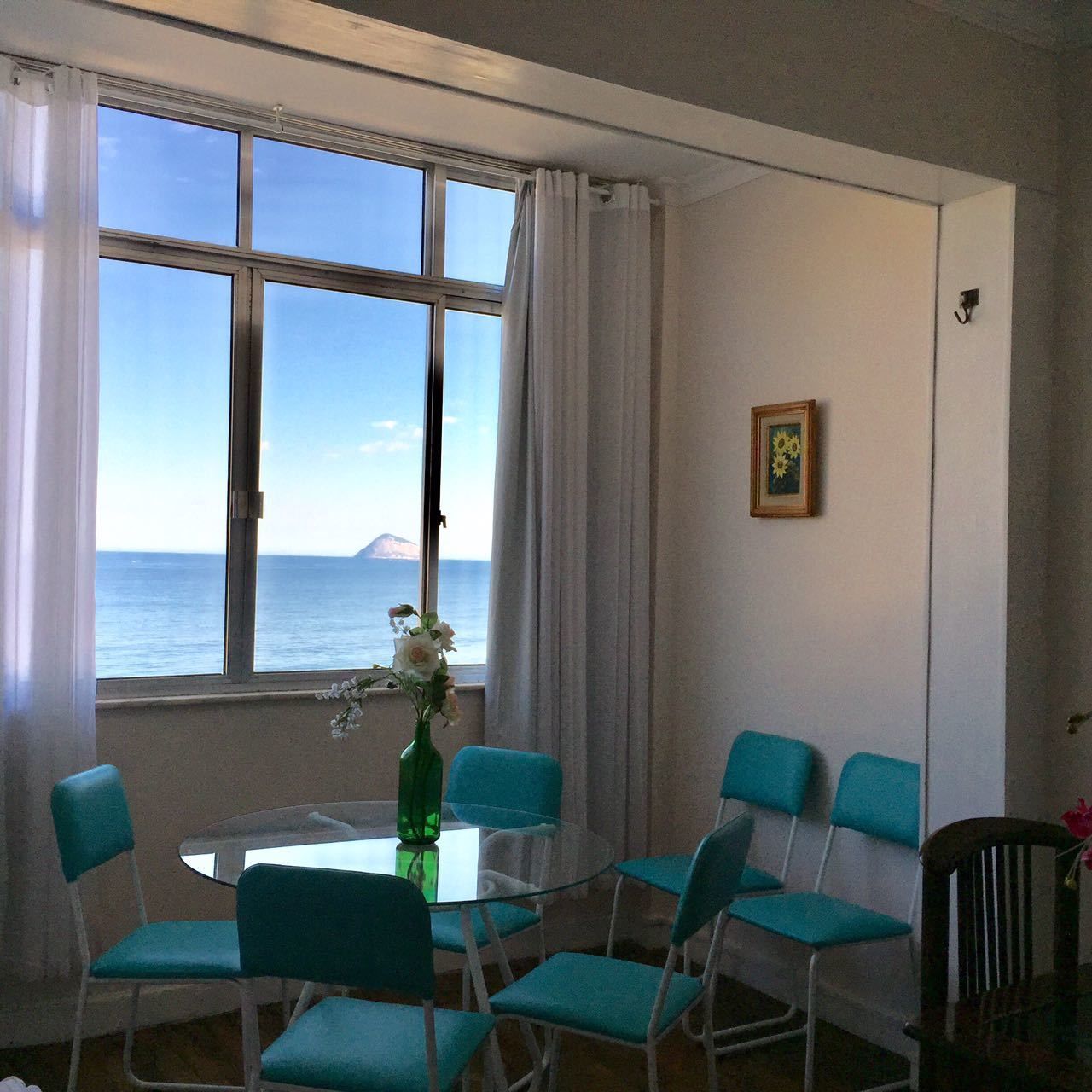 Een eetgedeelte met een ronde glazen tafel, zes teal stoelen, een groene fles en een vaas met bloemen. Een raam biedt uitzicht op de zee en een berg in de verte.