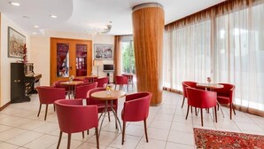 Un hall d’hôtel avec des fauteuils et des tables rouges, un grand piano et de grandes fenêtres ornées de rideaux légers.