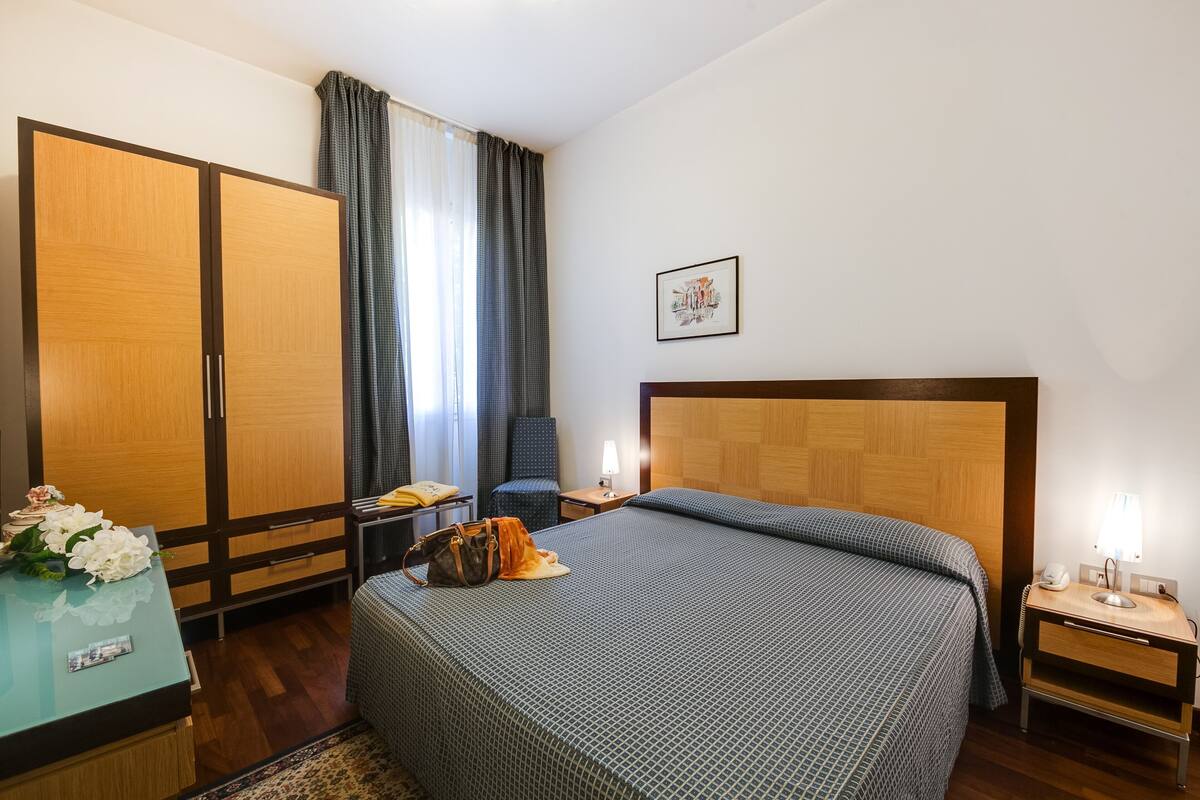 Une chambre d’hôtel comprenant un lit, un bureau, une chaise, une armoire et une table de chevet avec une lampe.
