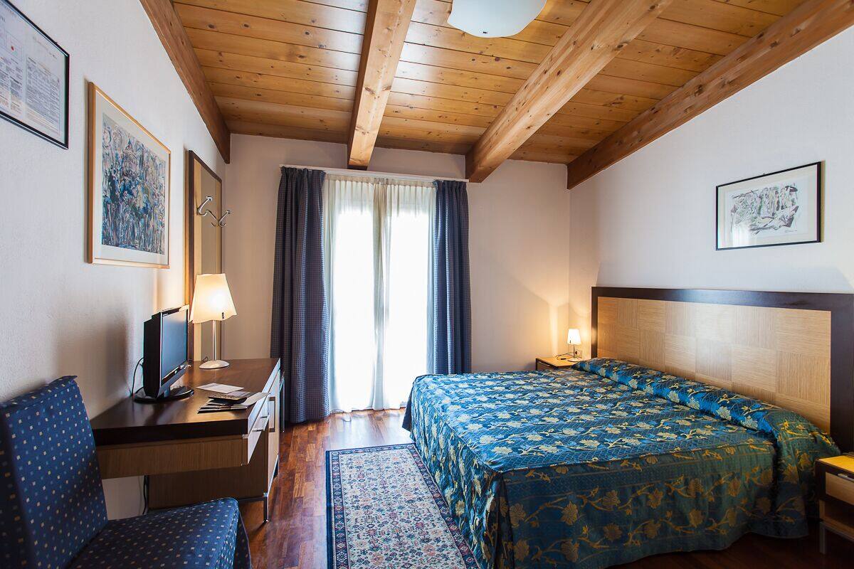 Une chambre d’hôtel avec un plafond en bois, un lit recouvert d’une couvre-lit à motifs floraux, un bureau avec un ordinateur, une chaise et une fenêtre avec des rideaux.