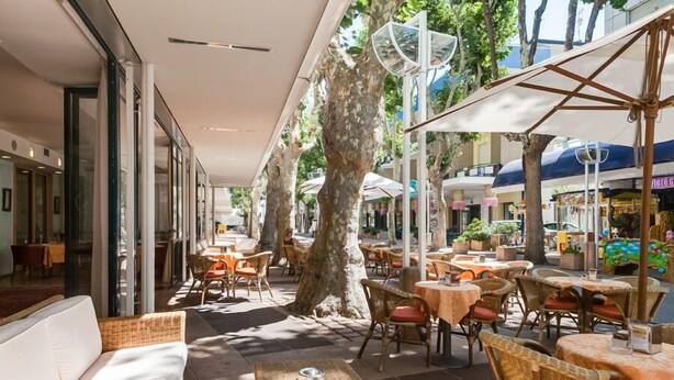 Un café en plein air avec des meubles en osier, des parasols et des arbres qui offrent de l’ombre.