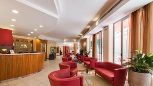 Un hall d’hôtel avec un comptoir d’accueil, un espace détente avec des sièges rouges et de grandes fenêtres.