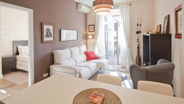 Un salon moderne avec un canapé blanc, une table à manger et un fauteuil gris. Il y a une grande fenêtre avec des rideaux, un luminaire au plafond et un téléviseur fixé au mur.
