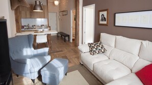 Un salon moderne avec un canapé blanc, un fauteuil bleu et un pouf assorti. On aperçoit, au fond, une cuisine avec des poutres en bois et un luminaire suspendu.