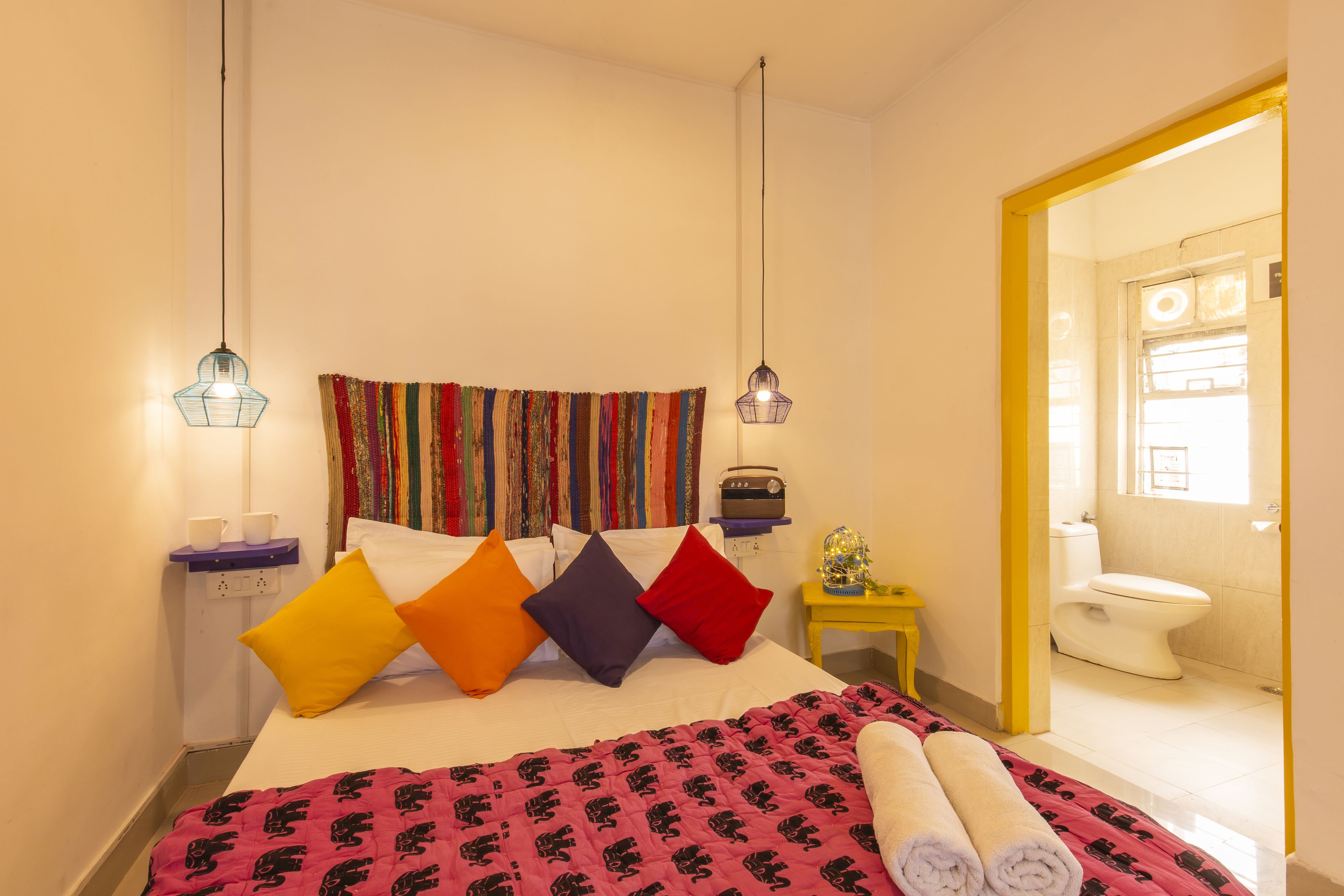 A bedroom with a bed, colorful pillows, a patterned bedspread, and a rolled towel.
