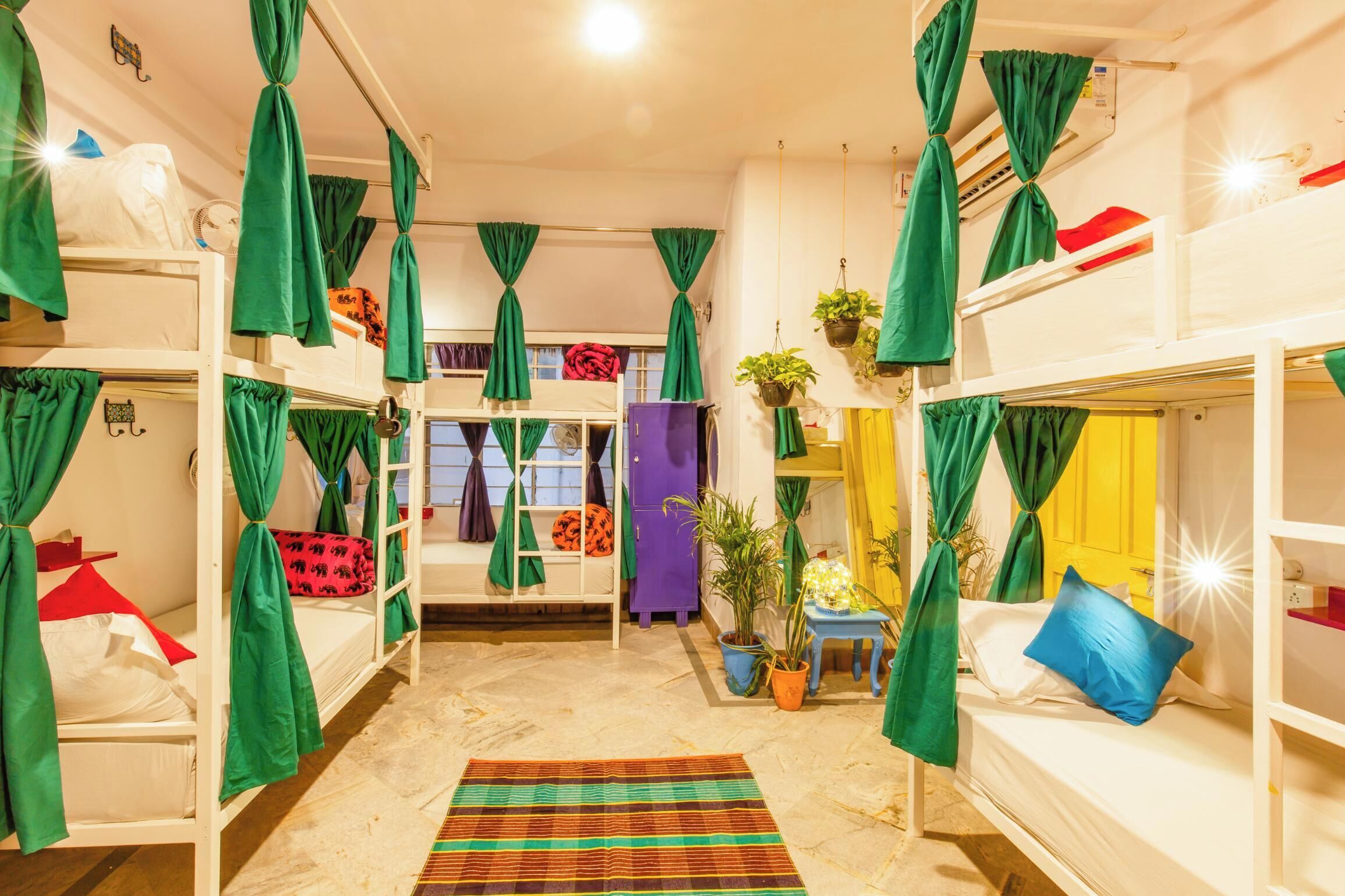 A dorm-style room with bunk beds, green curtains, potted plants, and a striped rug.