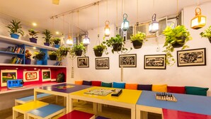 Een kleurrijke kamer met een lange tafel, bankjes, hangplanten en kunstwerken in lijsten aan de muren.