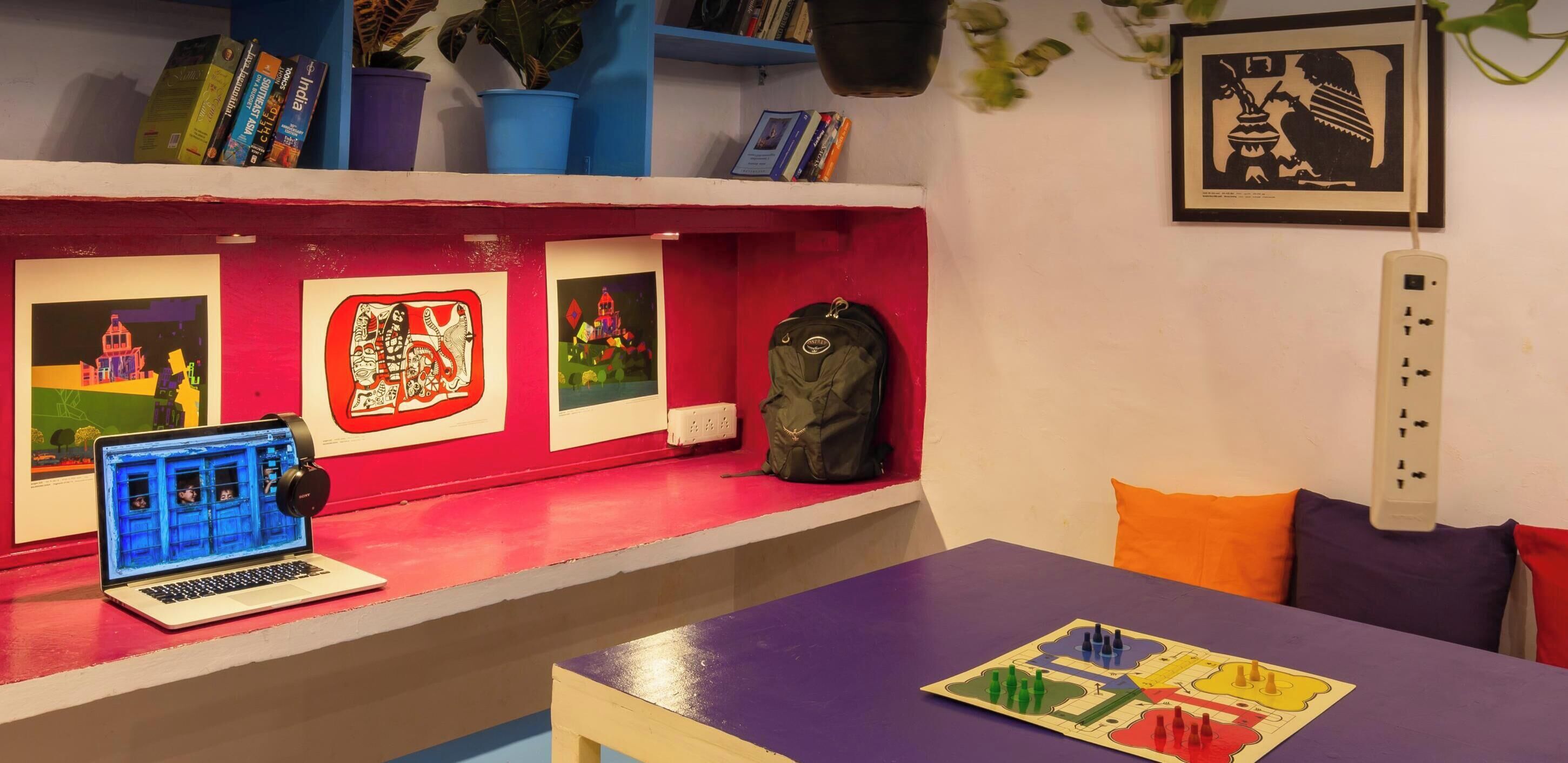 A room with a designated workstation featuring a laptop and headphones, surrounded by books and artwork on shelves, and a table with a board game.