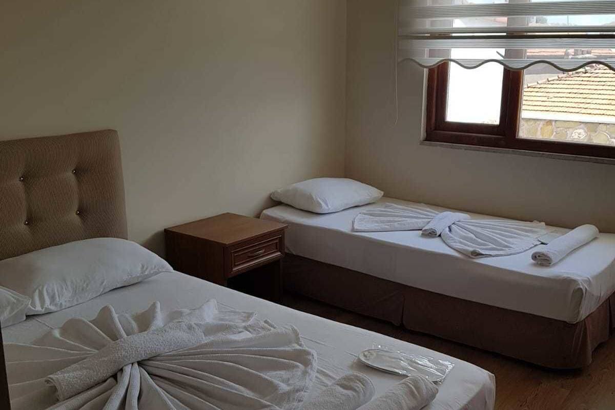 A hotel room with two beds, a wooden floor, a window with blinds, and a small bedside table.
