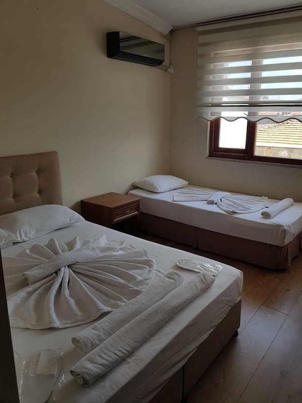A hotel room with two beds, a wooden floor, a window with blinds, and a small bedside table.