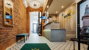 A hotel reception area with a brick wall, a wooden reception desk, a green carpet with the hotel name, and a statue on the wall.