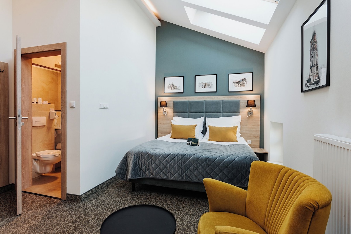Ein modernes Hotelzimmer mit Oberlicht, einem Bett mit grauer gesteppter Bettdecke, senffarbenen Kissen und einem gelben Sessel.