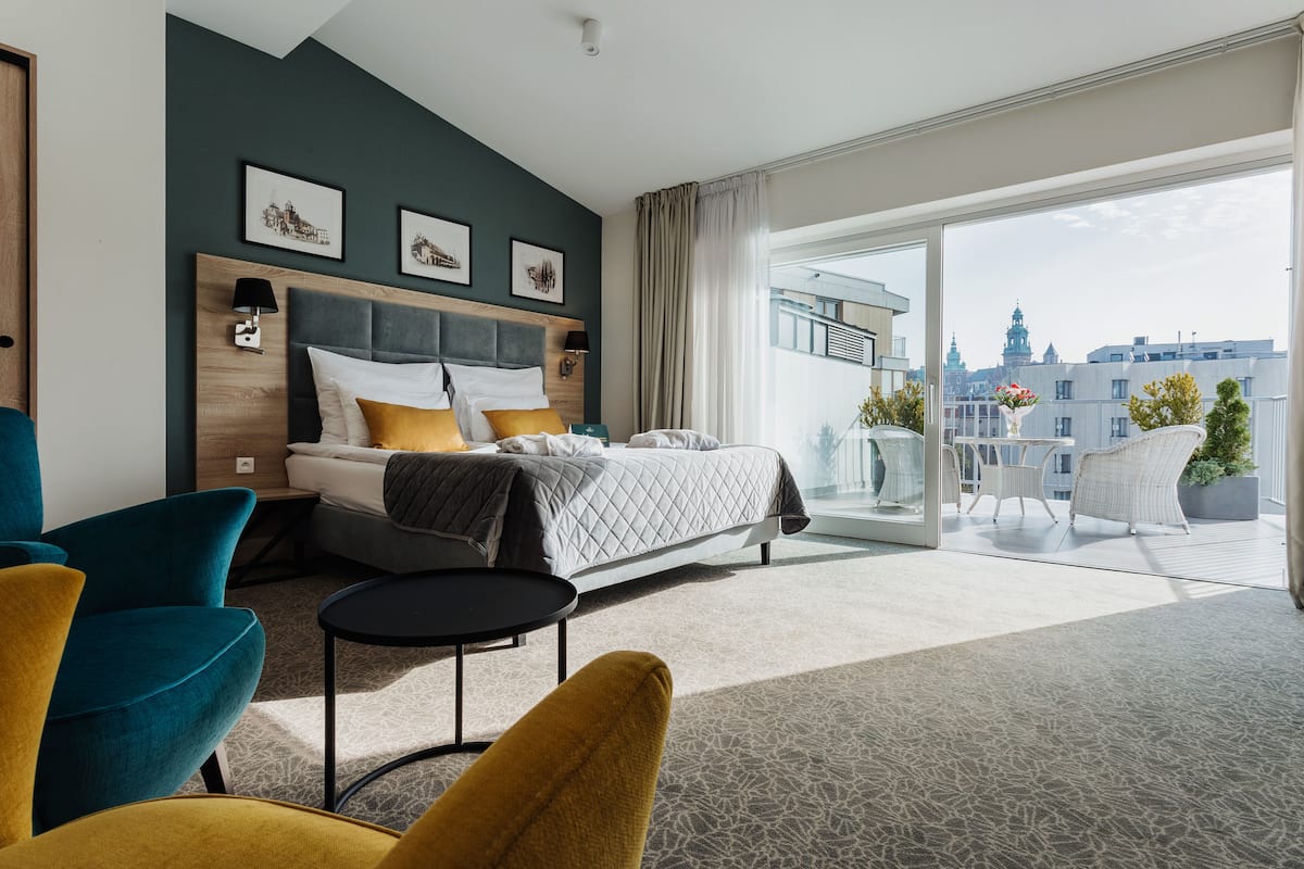 Une chambre d’hôtel moderne, équipée d’un lit, d’une petite table et de chaises. Elle dispose également d’un balcon avec vue sur la ville.