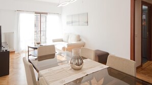 Een moderne woonkamer met een glazen eettafel, een bank en een salontafel. Een groot raam zorgt voor veel daglicht.