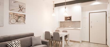 Un salon moderne avec un canapé gris, une table à manger avec des chaises et un coin cuisine doté d’un plan de travail blanc et de luminaires suspendus.