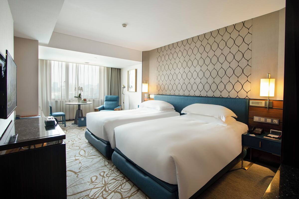 A hotel room with two beds, a desk, a chair, a TV, and a view of the city.