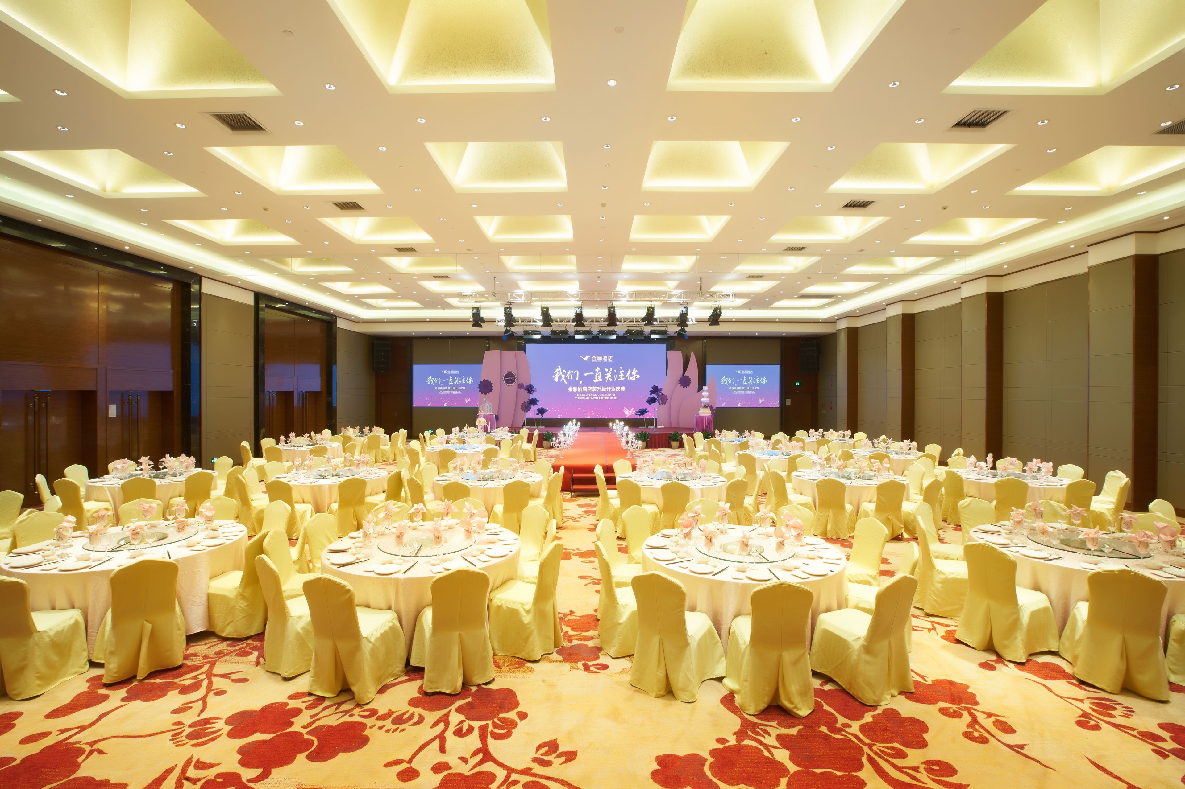 A large banquet hall with round tables set for events, a stage with a large screen, and a carpeted floor.