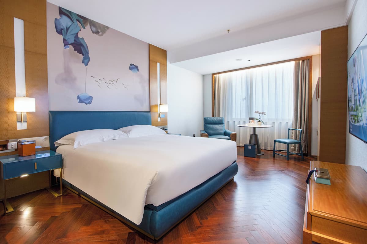 A hotel room with a large bed, a desk, a chair, a small table with a vase, and a painting on the wall.