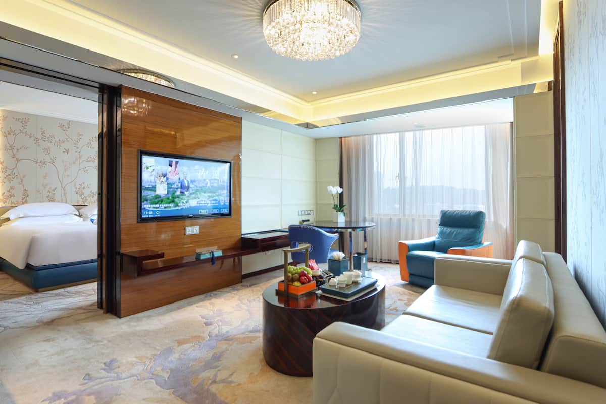 A hotel room with a bed, a TV, a seating area with a sofa and chairs, a coffee table with fruit, and a chandelier.
