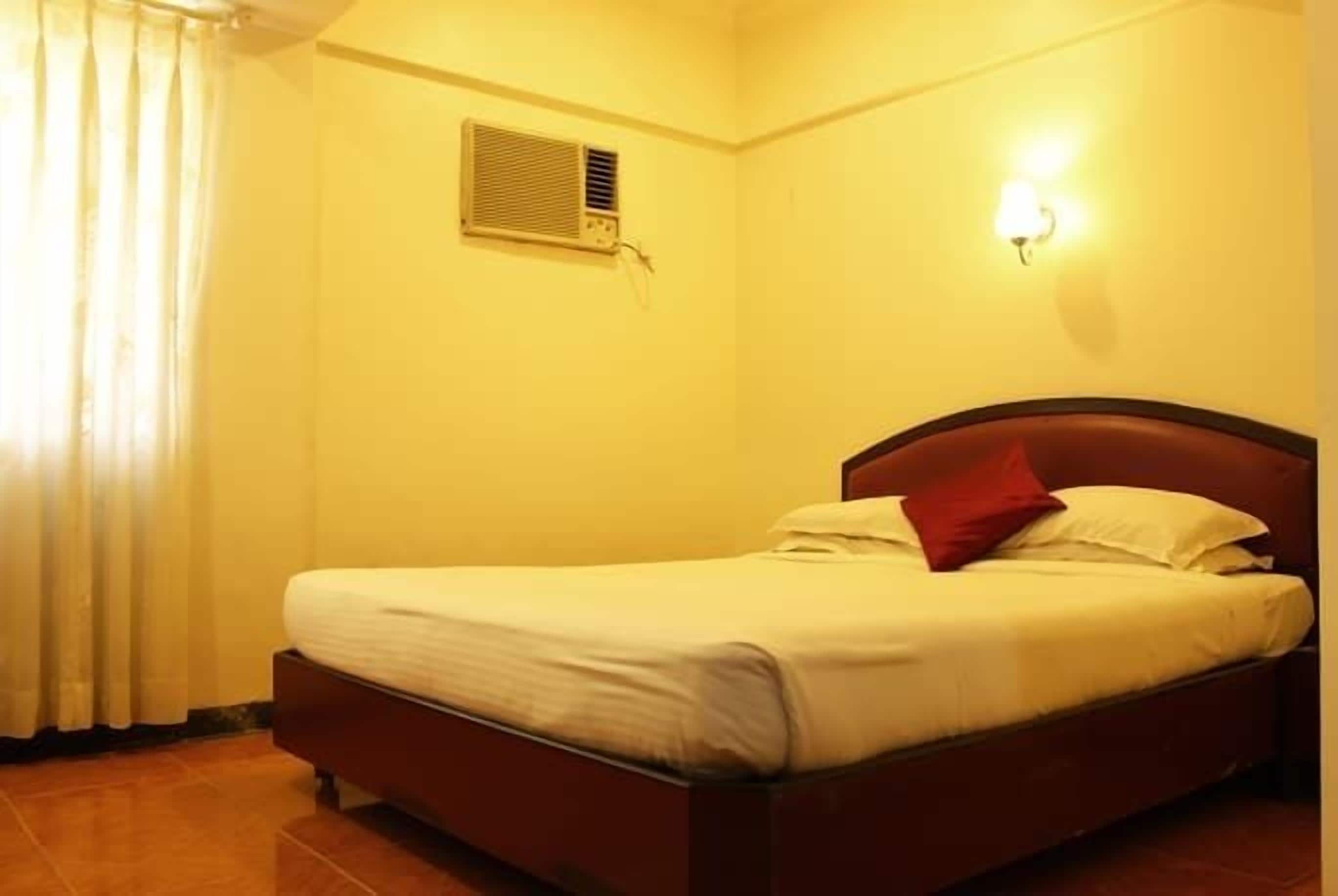 A single bed with a wooden headboard, a white mattress, and a red pillow. There is a wall-mounted air conditioning unit and a wall sconce.