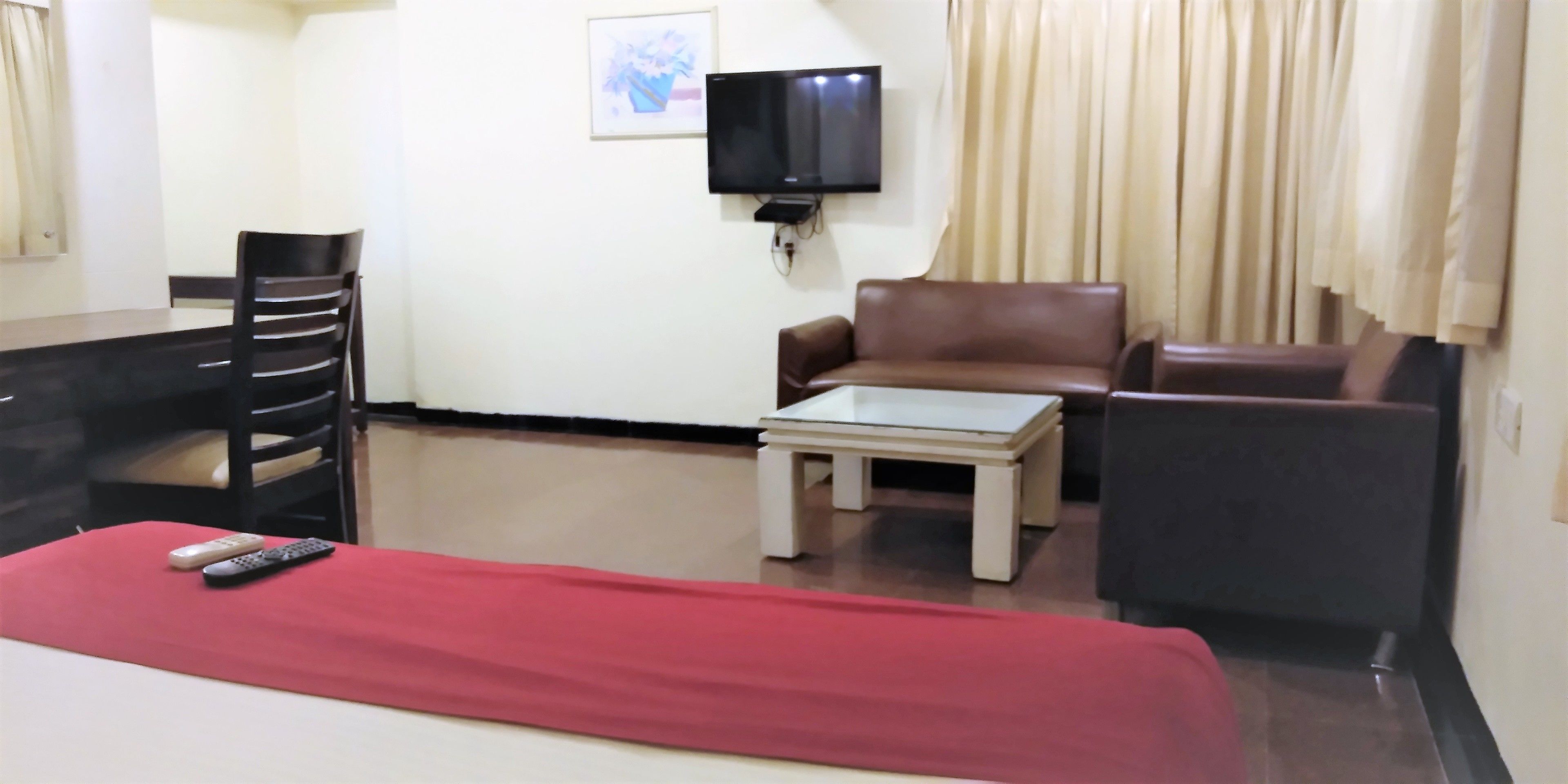 A hotel room with a bed, a red blanket, a brown leather sofa, a small white table, a TV mounted on the wall, and a chair.