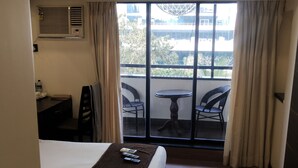 Quarto de hotel com cama, escrivaninha, duas cadeiras, mesa pequena e varanda com vista para edifícios e árvores.