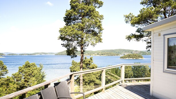 Una terraza de madera con sillas de mimbre que da a un lago, rodeada de árboles y un pequeño edificio con ventana.