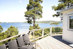 A wooden deck with wicker chairs overlooking a lake, surrounded by trees and a small building with a window. - Anfasteröd Gårdsvik (Ljungskile)