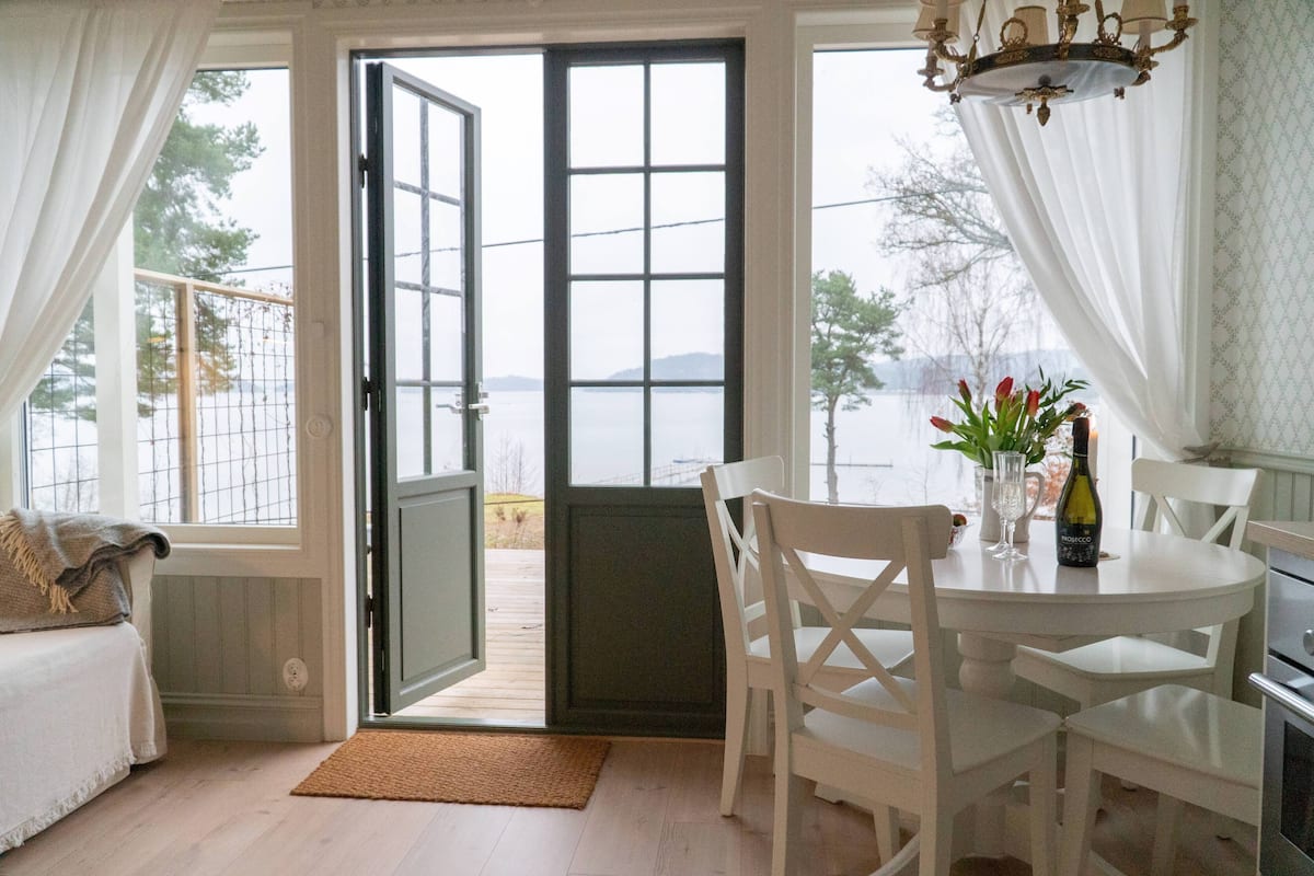 A room with a view of a body of water, a dining table with chairs, a chandelier, and a bottle of wine on the table.