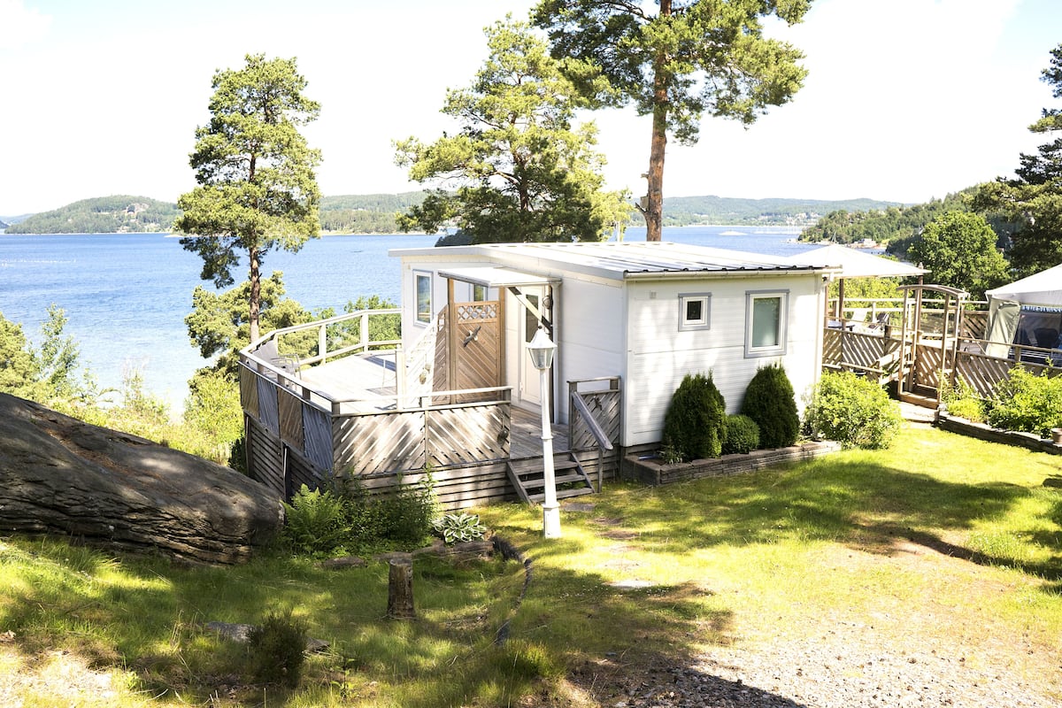 A small, single-story cabin with a deck overlooking a lake, surrounded by trees and a grassy yard.
