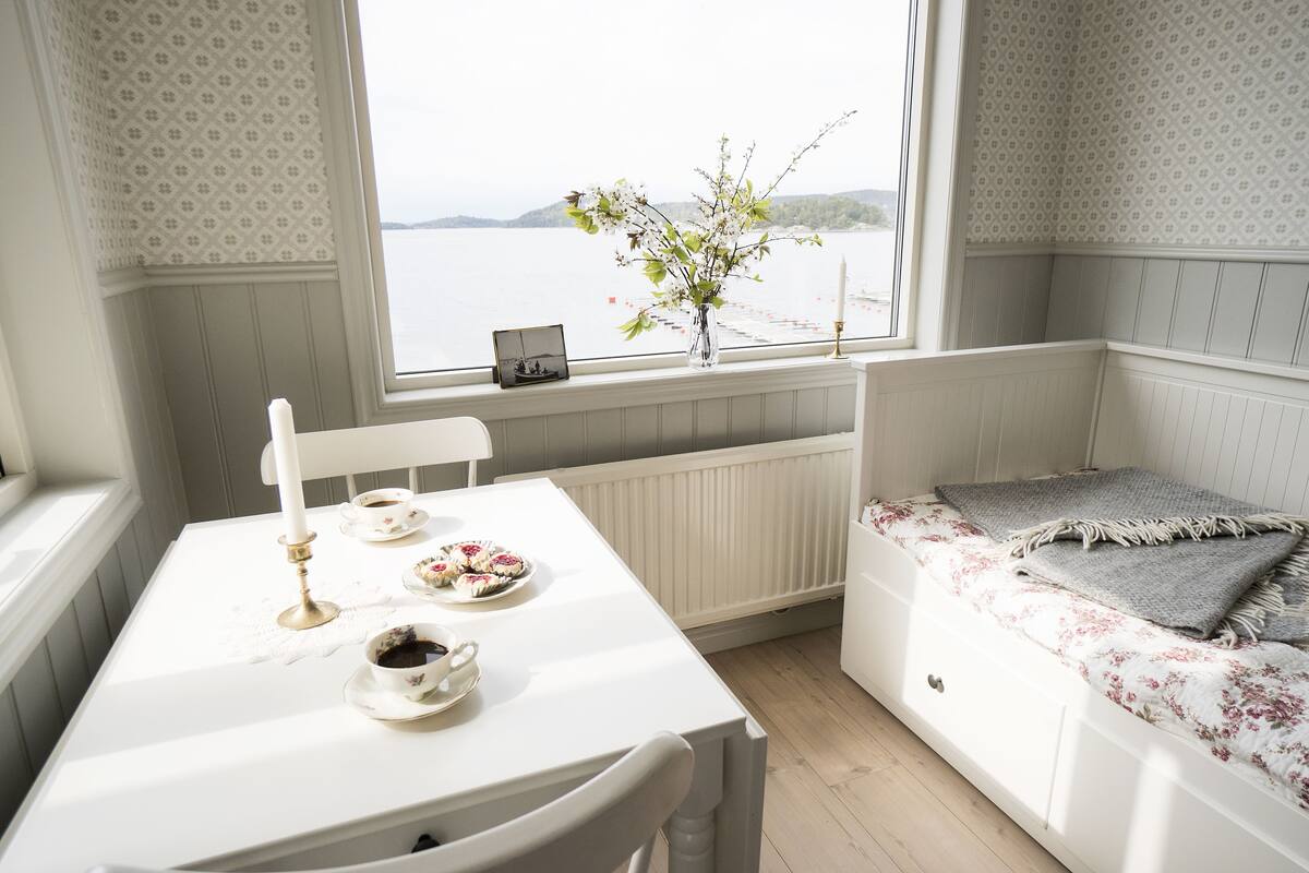 A room with a white table set for tea, a window with a view of the water, and a bed with a floral bedspread.