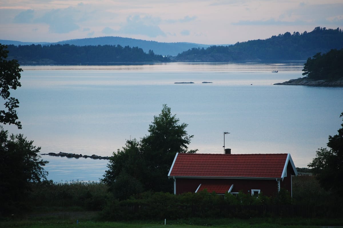 A red-roofed building with a white window, surrounded by greenery, overlooking a calm body of water with distant hills.