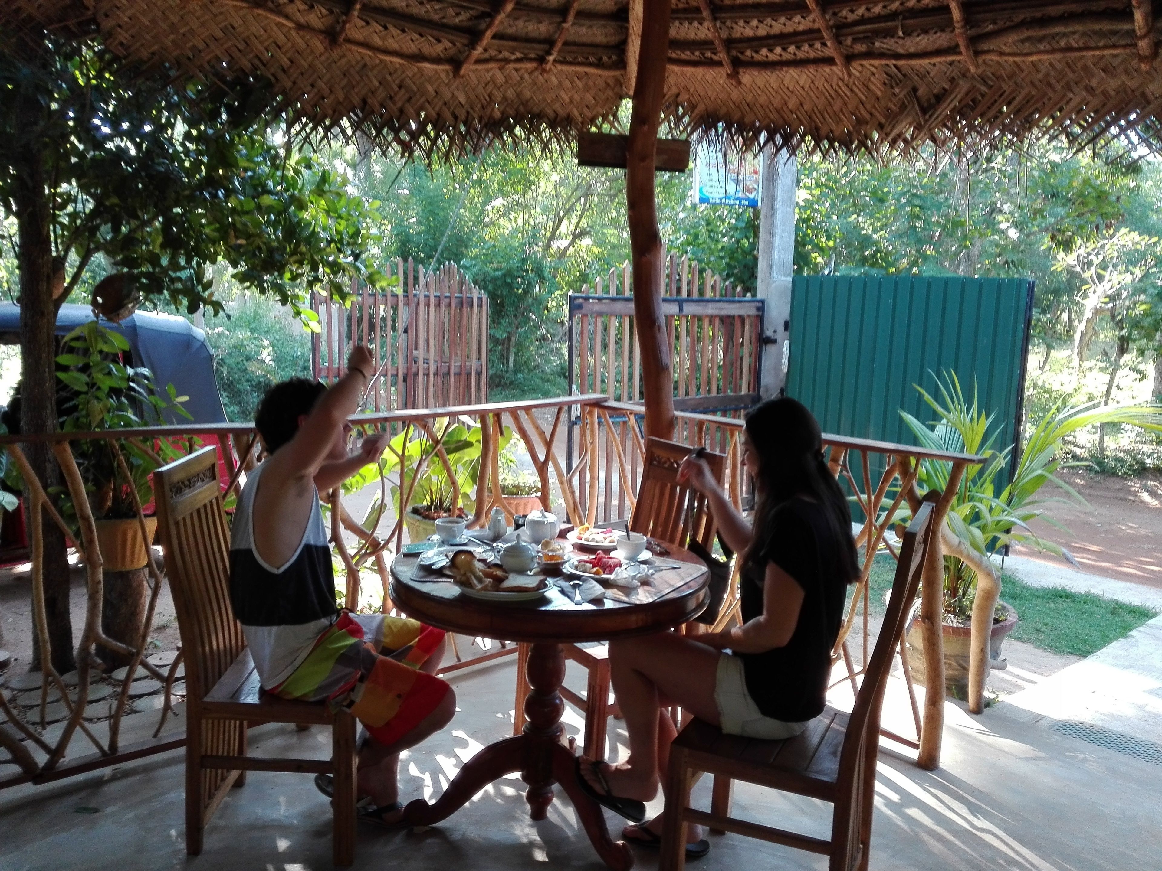 An outdoor dining area with a thatched roof, wooden chairs, and a table set for two.