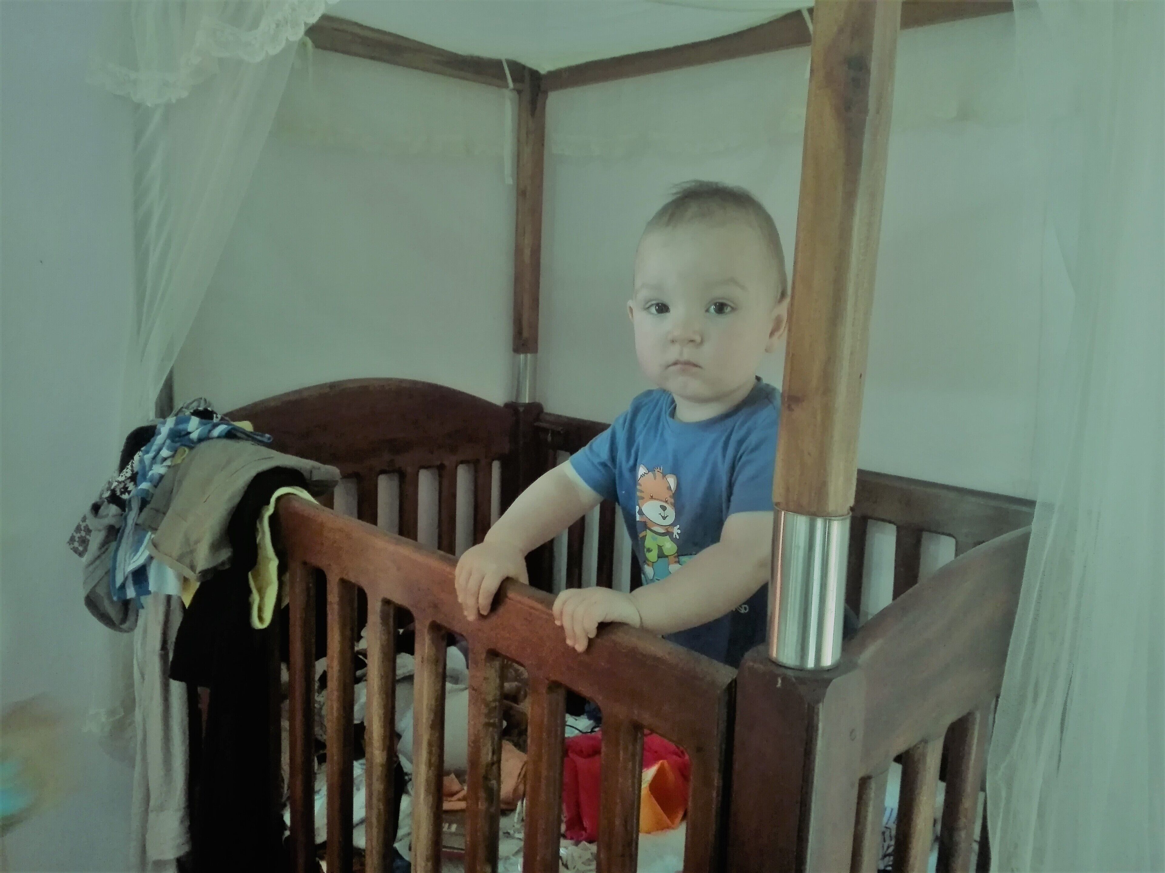 A child sitting in a wooden crib with a metal pole.
