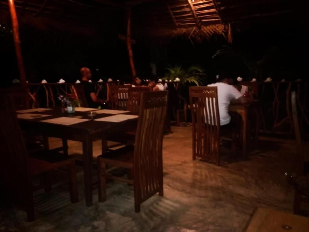Un espacio de comedor al aire libre con mesas y sillas de madera, un techo de paja y una palmera al fondo.