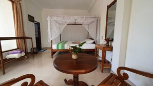 Une pièce avec un lit sous un dais blanc, une table ronde en bois ornée d’un vase de fleurs, une chaise en bois et une petite étagère en bois sur laquelle se trouvent une bouteille et une plante.