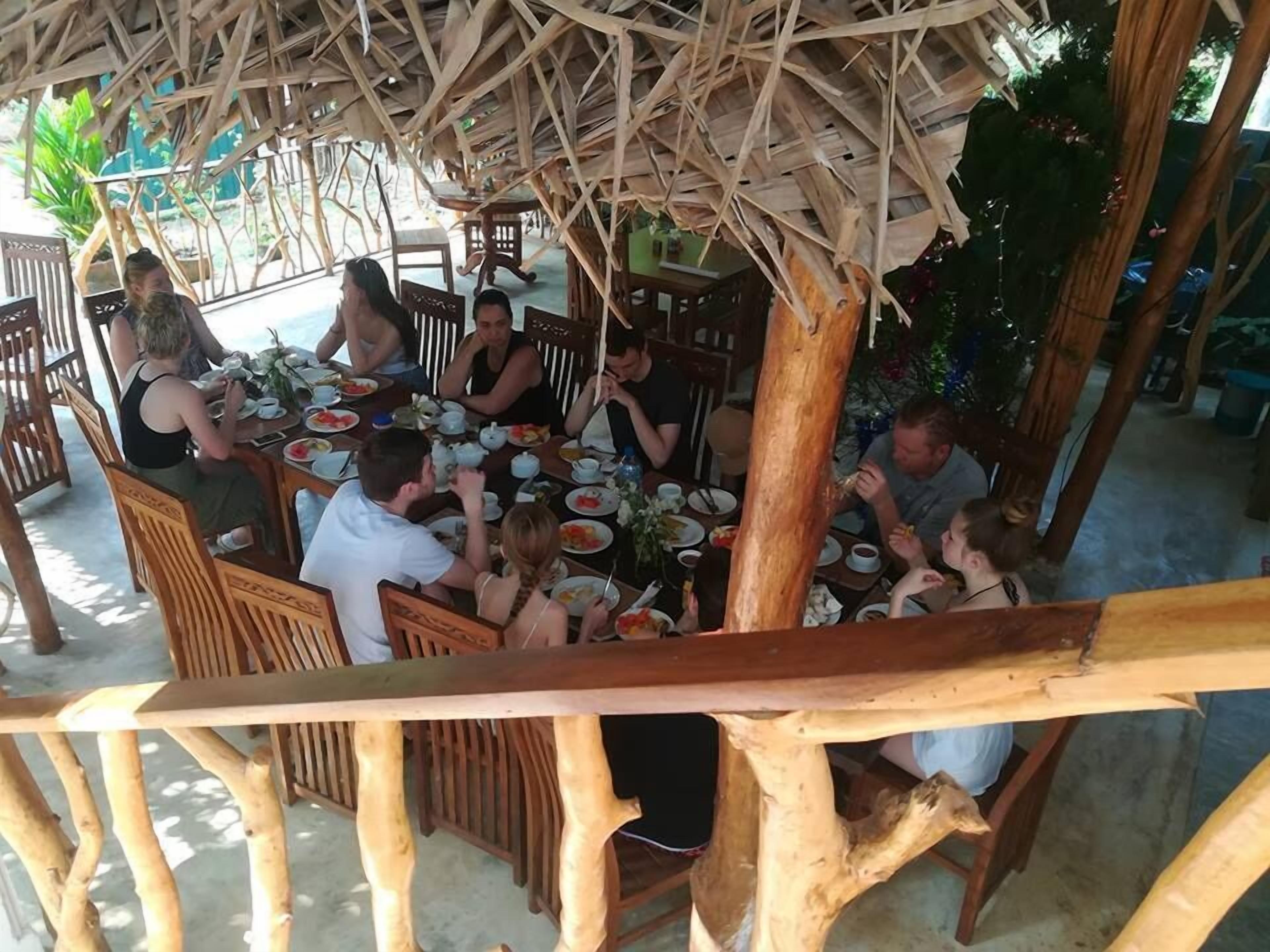 A group of people dining under a thatched roof structure with wooden furniture.