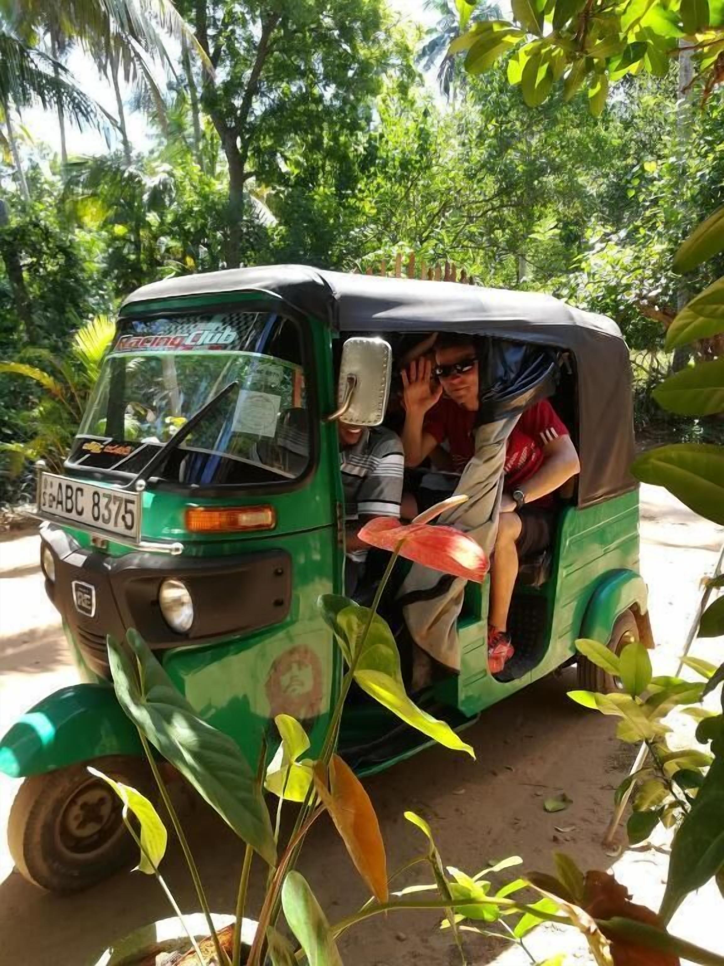 Un tuk-tuk vert, avec une partie arrière fermée, est garé dans un cadre naturel, entouré d’arbres et de plantes.