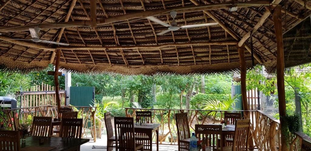 An outdoor dining area with wooden tables and chairs, a thatched roof, and lush greenery in the background.