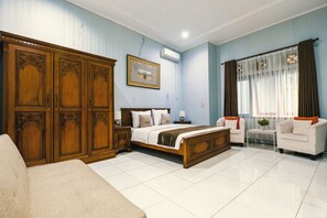 A hotel room with a large wooden wardrobe, a bed with white bedding, a small table, a white armchair, and a window with curtains. - Aleyra Hotel and Villa Garut (Garut)