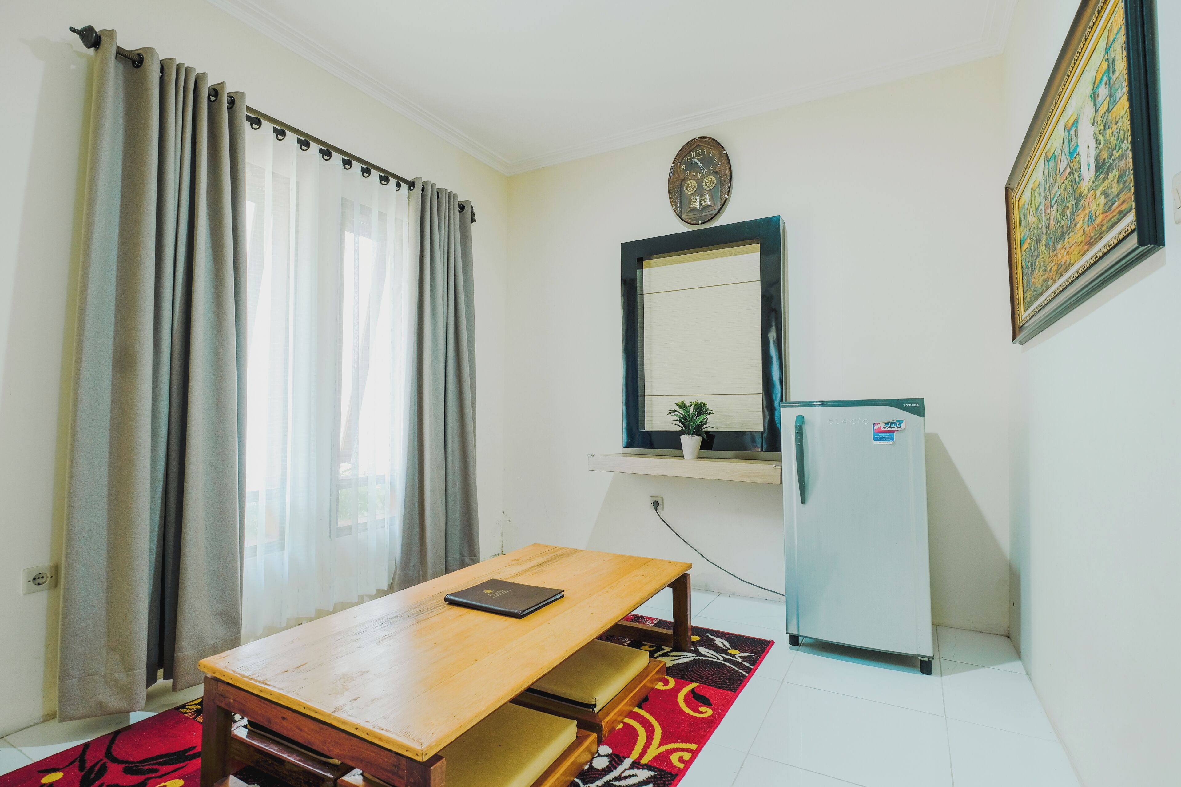 A small room with a wooden table, a refrigerator, a mirror, and a window with curtains.
