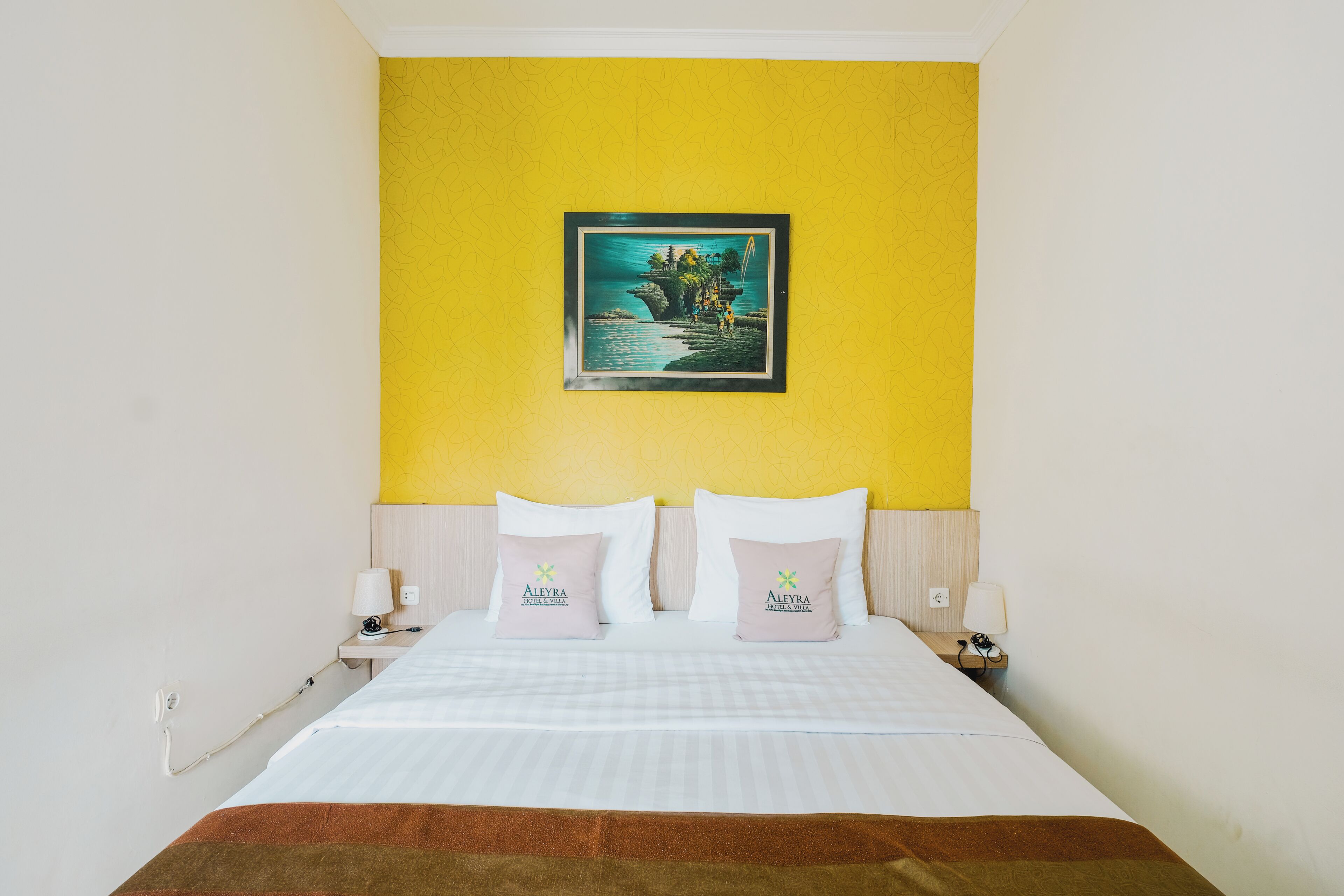 Habitación de hotel con cama, dos almohadas, mesita de noche, lámpara y un cuadro en la pared.