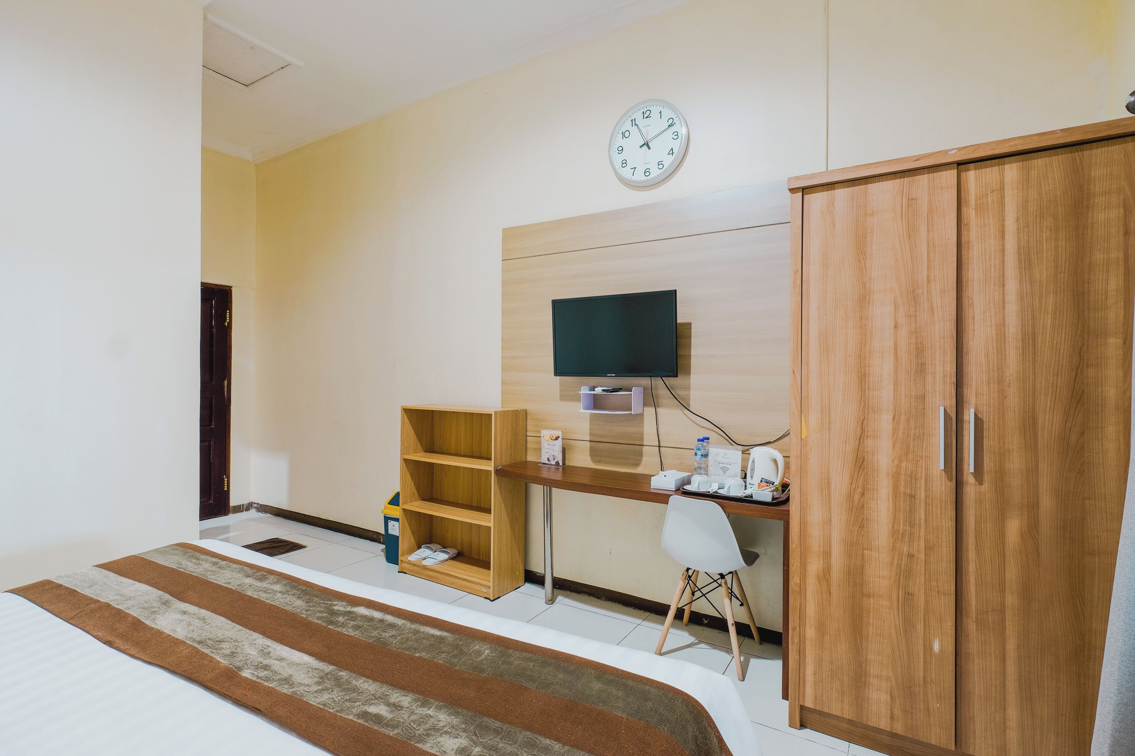A hotel room with a bed, a wooden desk with a TV, a wardrobe, and a small shelf with toiletries.