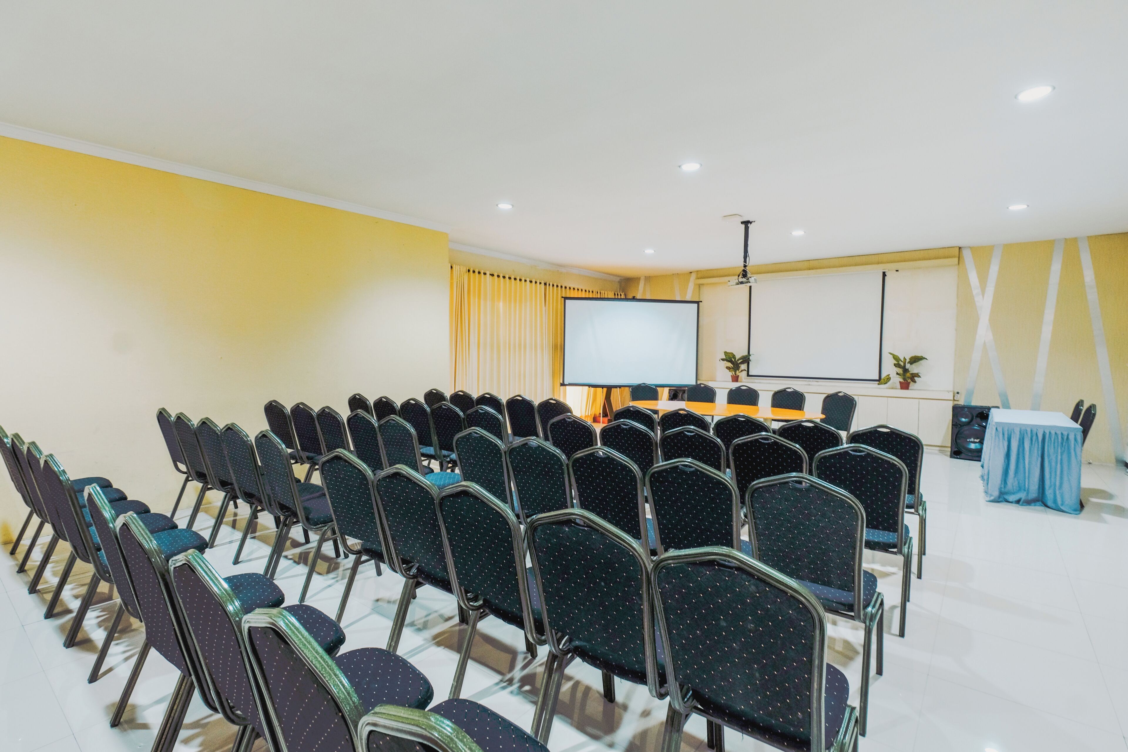 A conference room with rows of chairs facing a projection screen, a table with a blue tablecloth, and a speaker system.