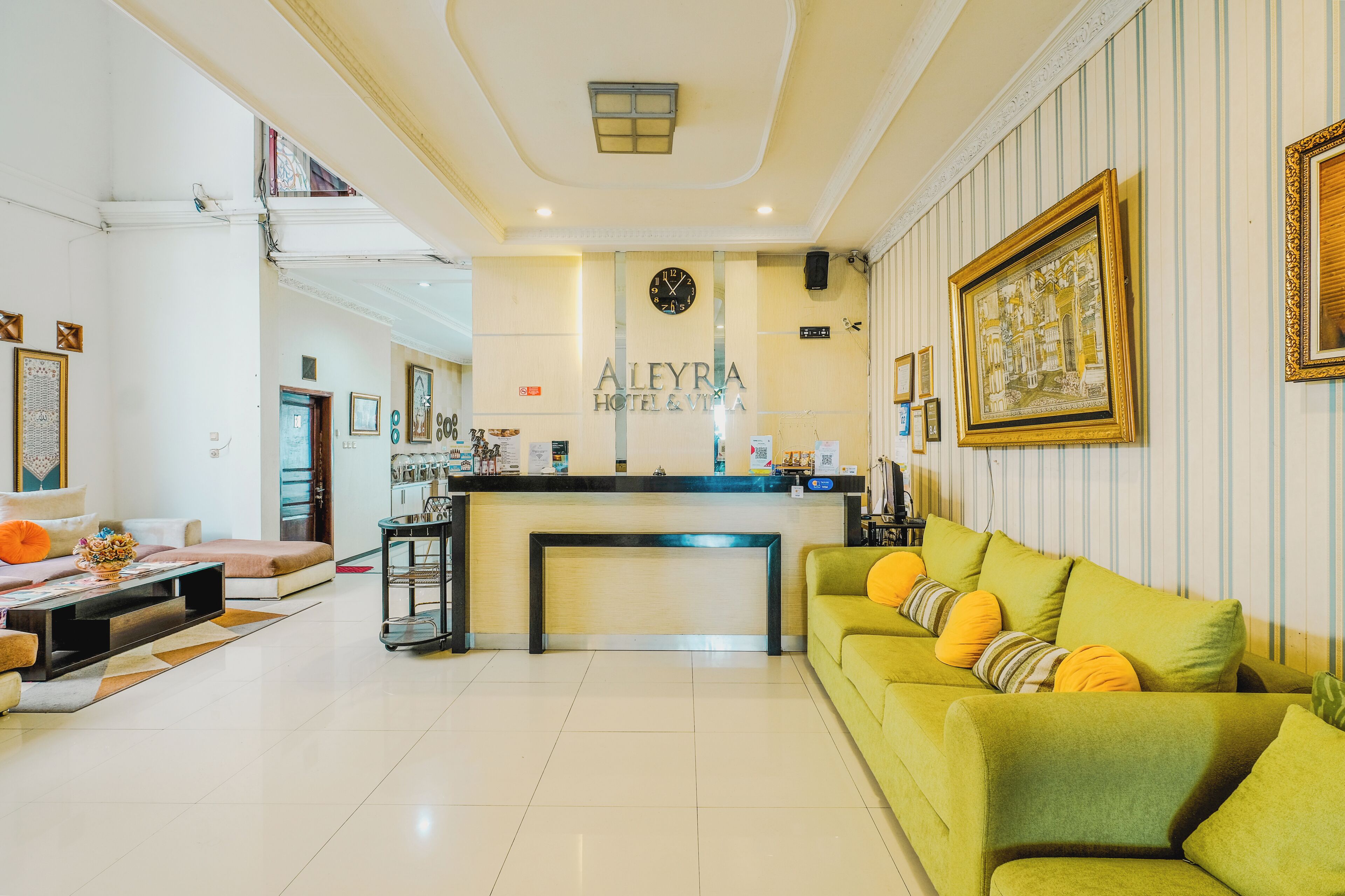A hotel lobby with a reception desk, a green sofa with cushions, a clock, and a framed picture on the wall.