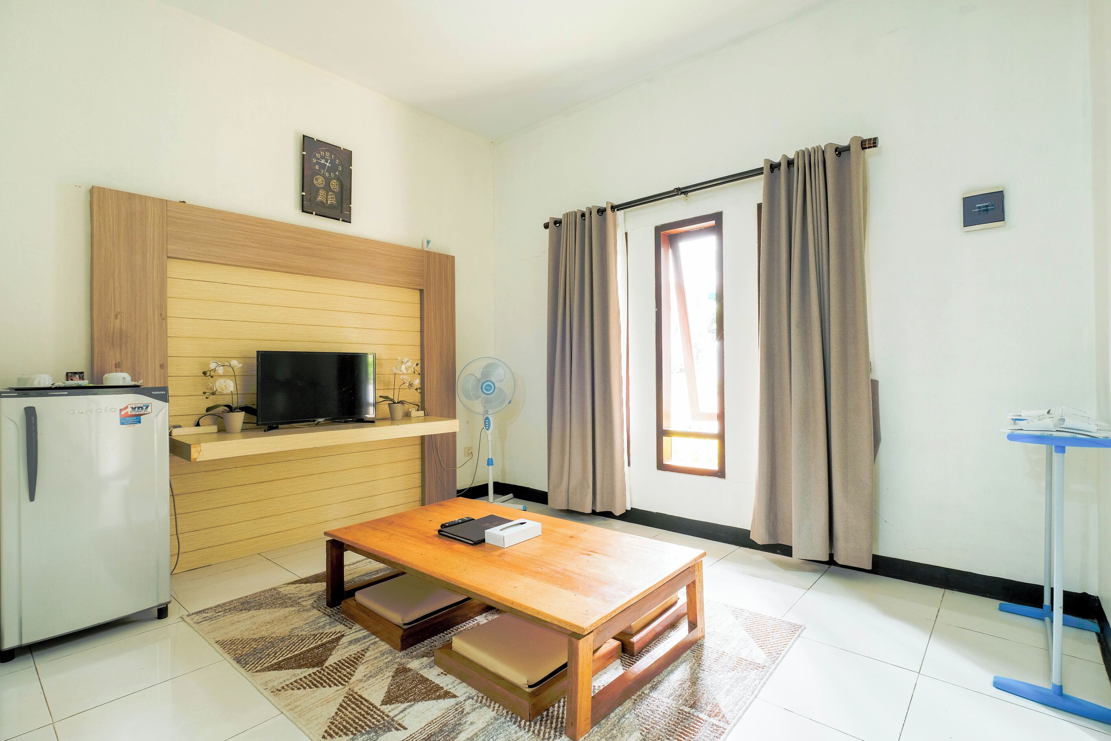 A compact living space with a kitchenette, a flat-screen TV, a wooden table, a small refrigerator, a standing fan, and a window with curtains.