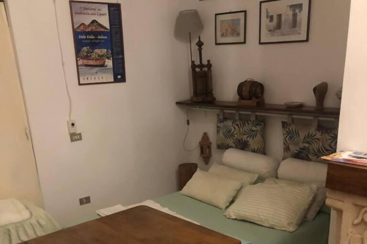 Une petite pièce accueillante, avec un lit, une table en bois et un sol carrelé. Des tableaux encadrés ornent le mur et une lampe est posée sur une étagère.