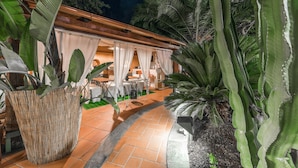 Een buitenterras met tafels en stoelen, omgeven door tropische planten en een houten pergola.