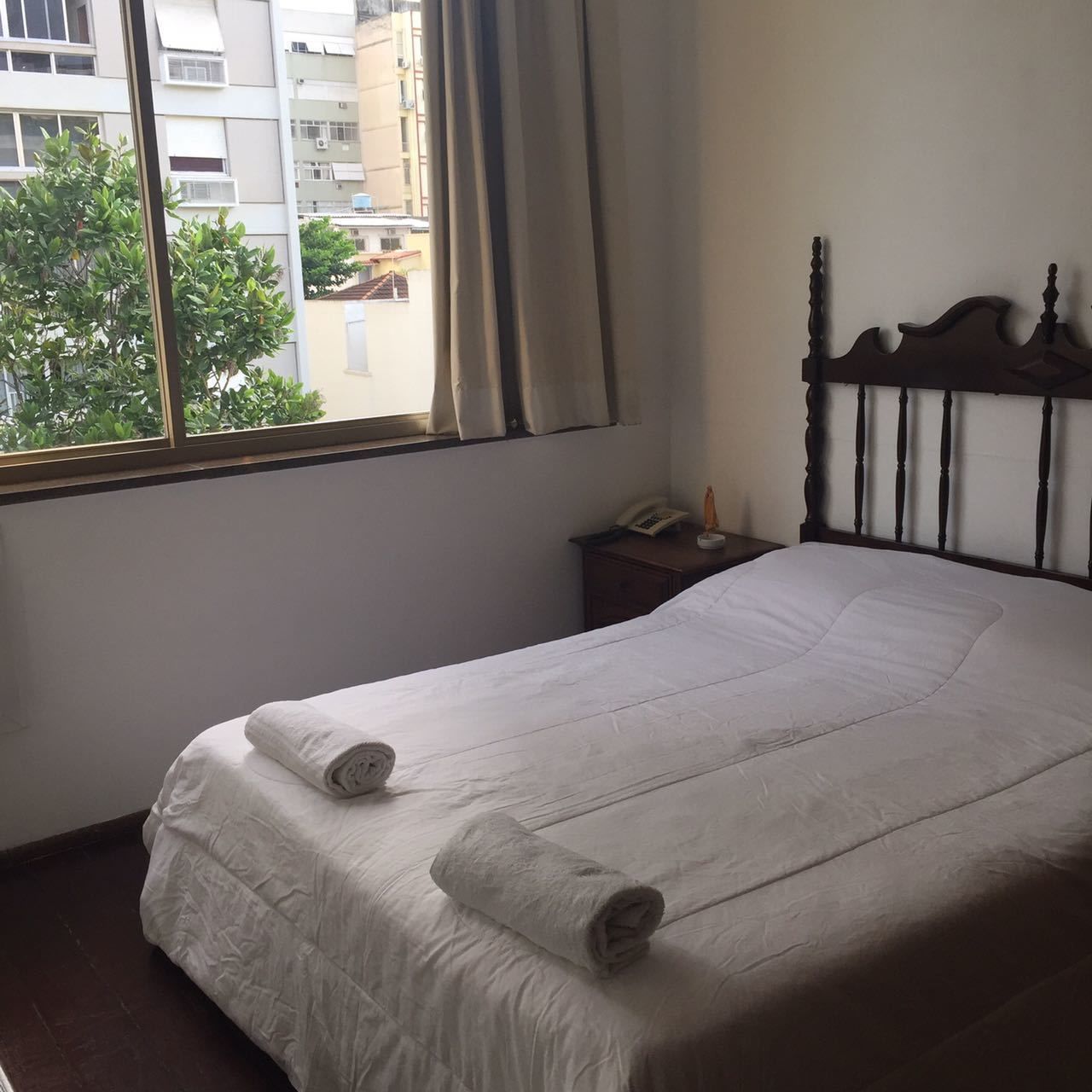 Una cama bien hecha con sábanas blancas y dos toallas enrolladas, una mesita de noche con una lámpara, y una ventana con cortinas.