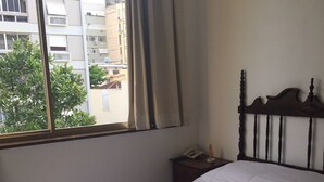 Una cama bien hecha con sábanas blancas y dos toallas enrolladas, una mesita de noche con una lámpara, y una ventana con cortinas.